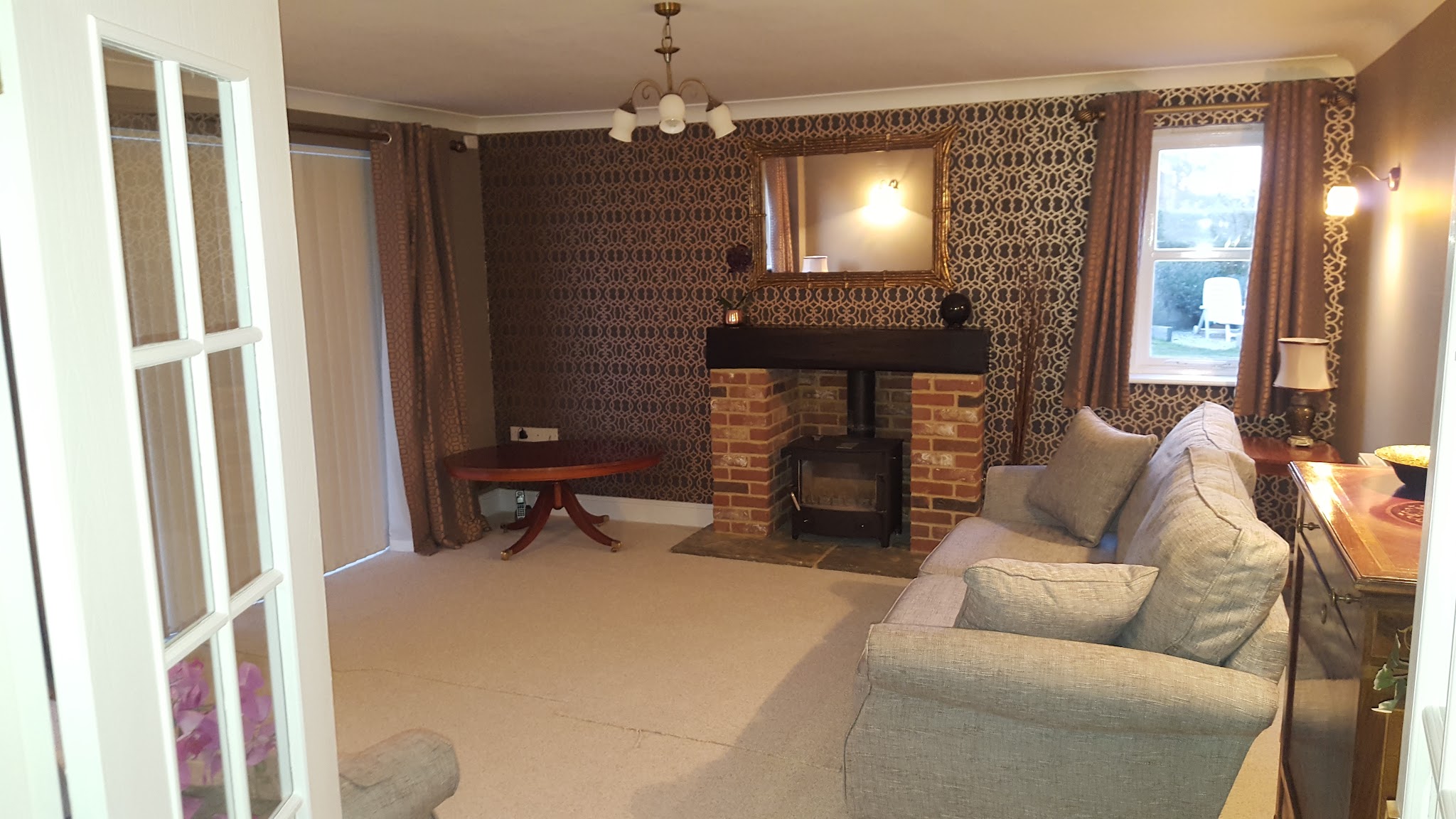 Living room with patterned feature wallpaper around a brick fireplace and wood burner